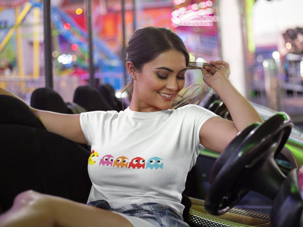 Woman sitting in a brightly lit arcade or carnival setting, wearing a white t-shirt featuring a pixelated design of Ms. Pac-Man and four colorful ghosts (pink, orange, red, and blue) arranged in a row. The retro design adds a playful, nostalgic touch to her casual outfit, paired with a plaid shirt tied at her waist. The background includes blurred neon lights and arcade rides, enhancing the fun, vibrant atmosphere.