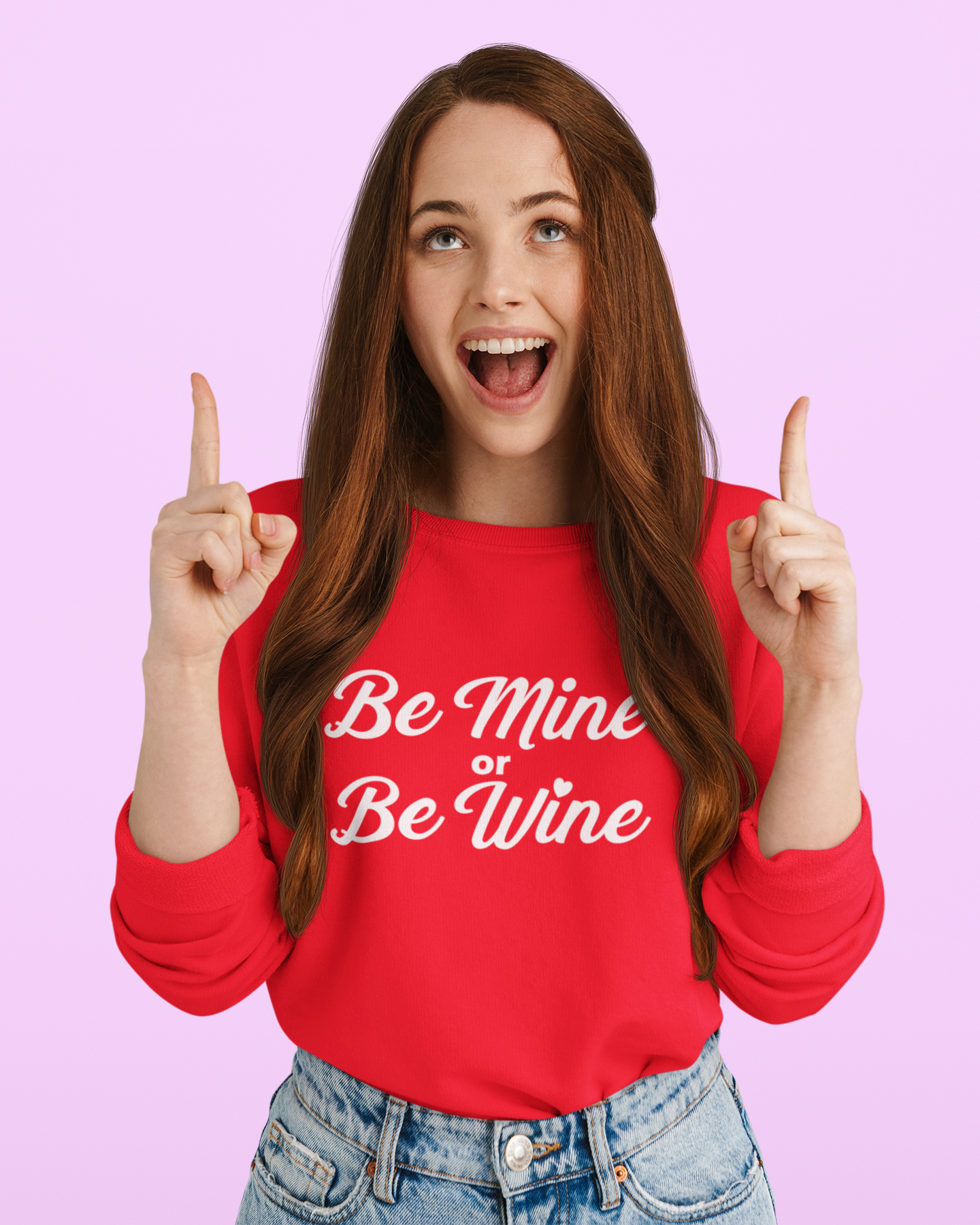 A cheerful young woman with long red hair, wearing a red sweatshirt that says "Be Mine or Be Wine" in white script font, smiles widely while pointing upward with both hands. The background is a soft pastel pink.