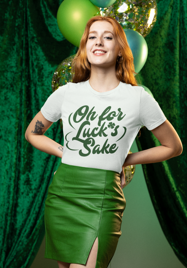 A red-haired woman with a confident smile poses in a festive green setting with balloons and shimmering decorations in the background. She wears a white t-shirt featuring the phrase "Oh for Luck's Sake" in stylish green script, paired with a high-waisted green leather skirt with a front slit. Her arms are on her hips, and she has a tattoo on her left arm. The overall scene exudes a St. Patrick's Day theme with vibrant green hues.