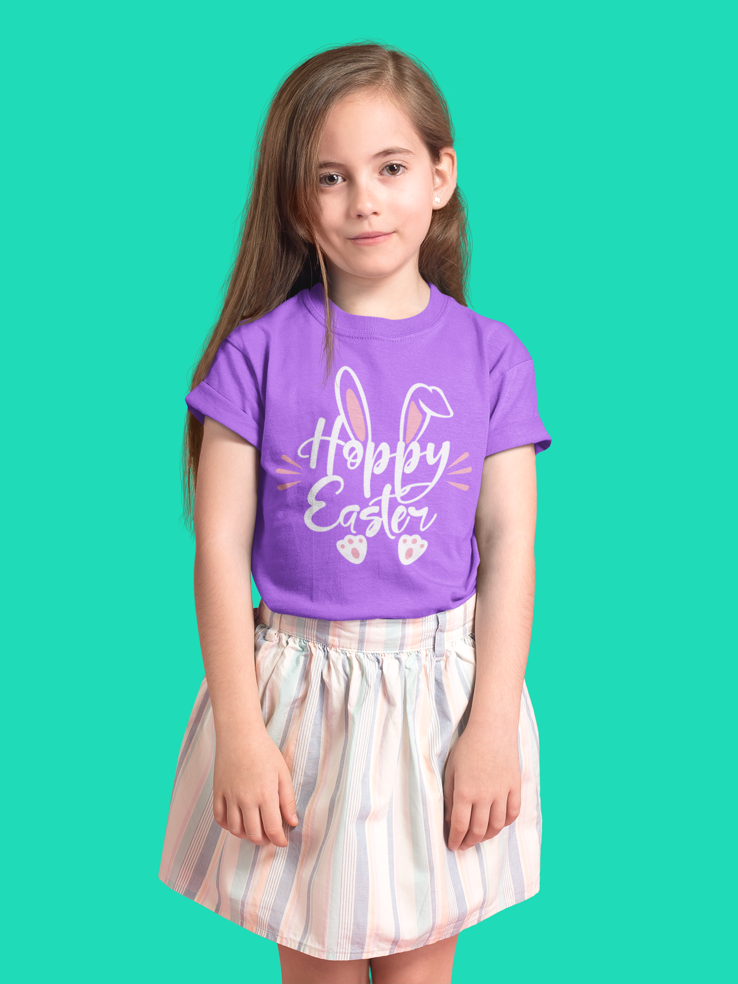 Young girl with long brown hair wearing a purple t-shirt that says "Hoppy Easter" with bunny ears, pink accents, and paw prints. She is standing against a teal background and wearing a light-colored striped skirt.